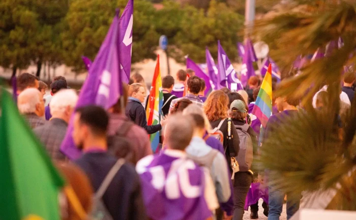 Volt Malta supporters marching with purple flags and a pride flag at a political rally.